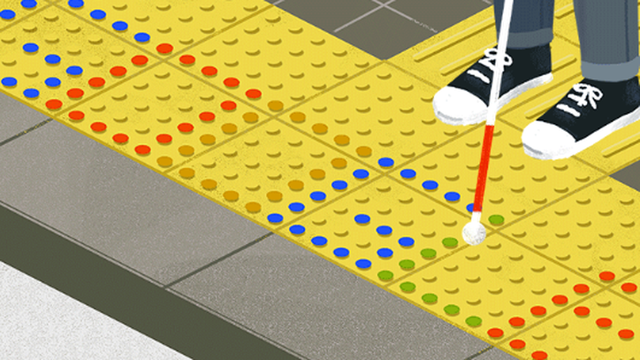 A person wearing black shoes stands with a red and white cane on yellow dotted tactile pavement. Some dots are different colors and spell out Google